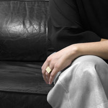 Load image into Gallery viewer, A person wearing the Brass Square Ring on their ring finger while sitting on a black leather sofa