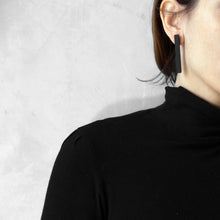 Load image into Gallery viewer, A woman wearing the Black Column Earrings while touching her hair with a black bluse on a grey background