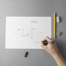 Load image into Gallery viewer, Design on paper of the Earring's project with a hand that draws, eraser and sharpener.