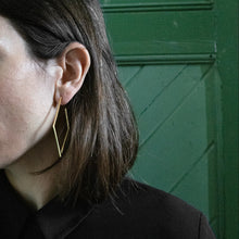 Laden Sie das Bild in den Galerie-Viewer, Minimalist gold frame earrings worn outdoors against a contemporary city backdrop with contrasting light and reflections.