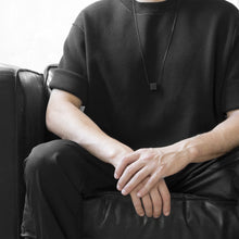Load image into Gallery viewer, A man wearing the Black MODULOUNO Necklace with a black t-shirt on a grey background while sitting on a black sofa