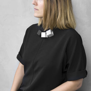 A woman with a blond hair wearing the Zero Elements Necklace with a black bluse on a grey background 
