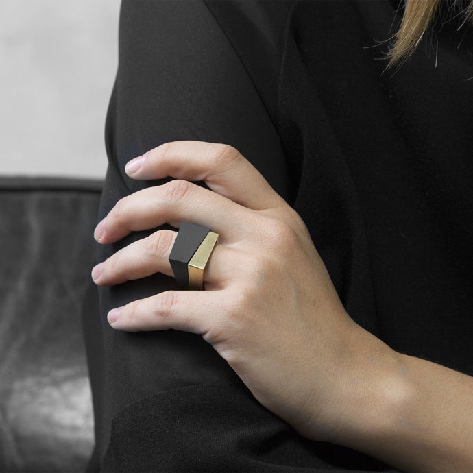 A woman wearing the Assembled Ring, a combination of MK3 black asymmetric ring and Brass squared ring, on their ring finger while sitting on a black leather sofa