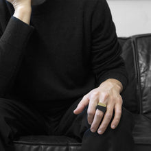 Load image into Gallery viewer, A man wearing the Assembled Ring, a combination of MK3 black asymmetric ring and Brass squared ring, on their ring finger while sitting on a black leather sofa